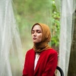 Elegant portrait of a woman wearing a red coat and hijab, sitting outdoors with closed eyes.